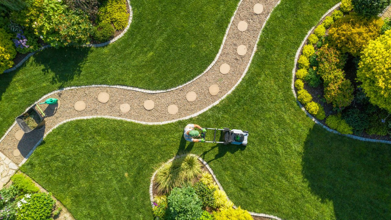 Aerial view of a landscaped garden with curved pathways and a person mowing a lush green lawn surrounded by plants and shrubs.