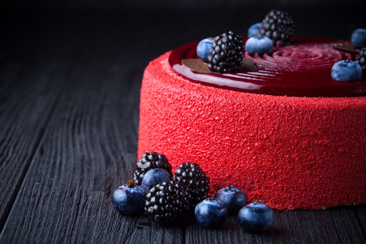 Red berry cake displayed on a dark wooden table with fresh blueberries and blackberries